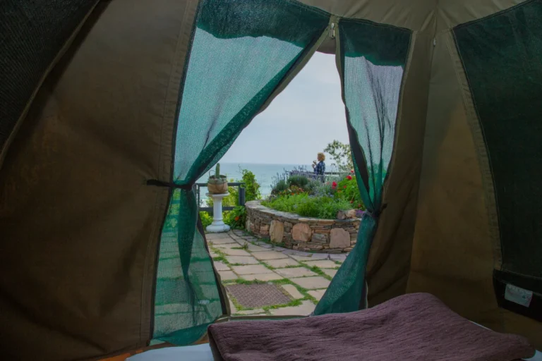 View through a garden tent doorway at Wilderness Beach House Backpackers, past a stone-walled flower bed on the terrace to the sea