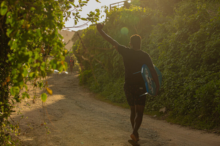 Guest carrying a surfboard up the leafy sand road to Wilderness Beach House Backpackers in golden evening light