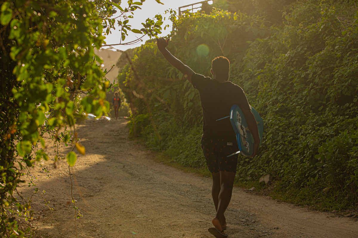 Guest carrying a surfboard up the leafy sand road to Wilderness Beach House Backpackers in golden evening light